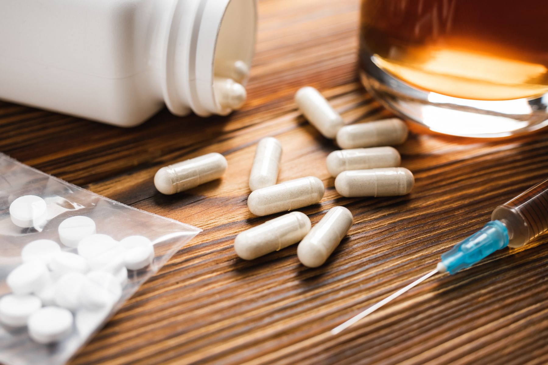 White pills, syringe with a drugs dose, narcotics, glass with alcohol drink on a wooden table, close-up view. Concept of addiction and bad habits